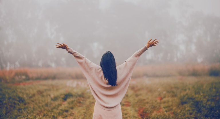 Femme de dos bras ouverts dans un champ brumeux, symbole de lâcher-prise